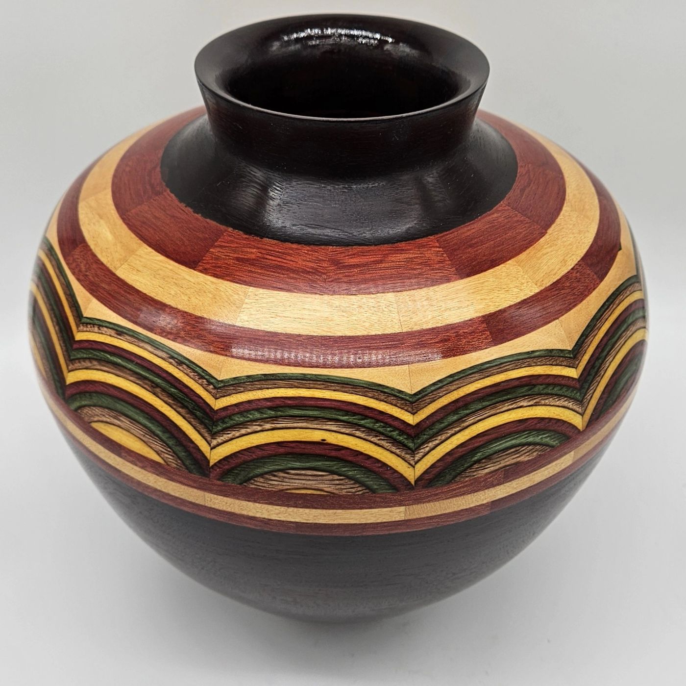 Globe-shaped segmented wooden vase with a dark collar above a wide band of concentric arcs in yellow, green, red and natural wood